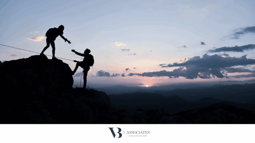 silhouette of 2 climbers against a blue/purple sky. one is helping the other reach the next height, holding their hand out.